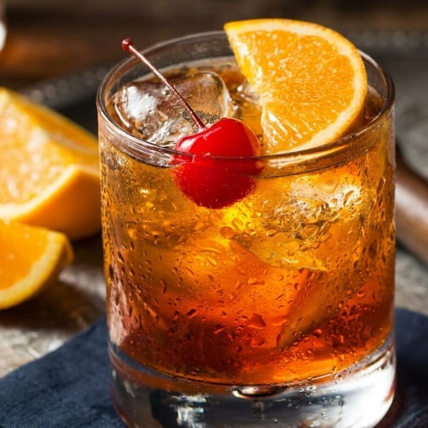 Old Fashioned cocktail with orange slice and cherry on a silver tray with orange wedges.