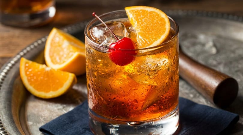 Old Fashioned cocktail with orange slice and cherry on a silver tray with orange wedges.