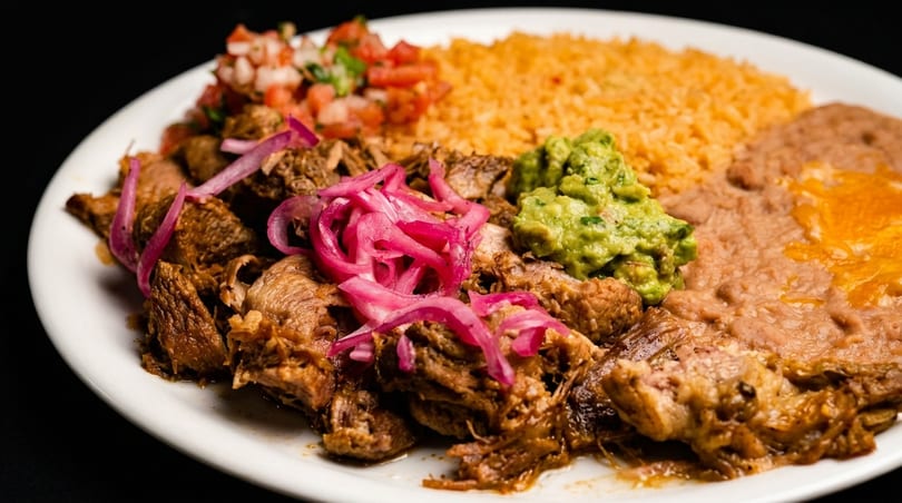 A plate of carnitas with pickled red onions, rice, refried beans, guacamole, and pico de gallo.