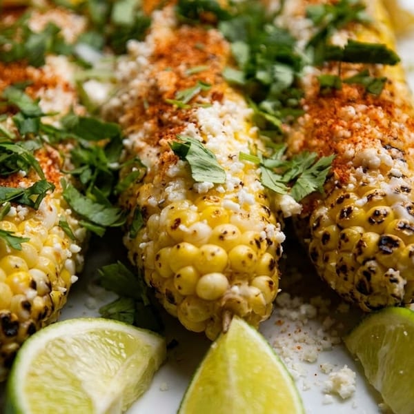 Three grilled elotes, Mexican street corn, topped with cheese, chili powder, cilantro, and lime wedges.