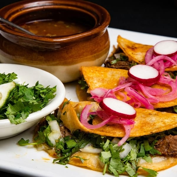 Three birria tacos with pickled red onions and radishes, served with consommé, cilantro, and lime.