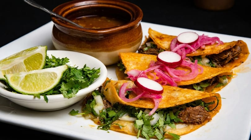 Three birria tacos with pickled red onions and radishes, served with consommé, cilantro, and lime.