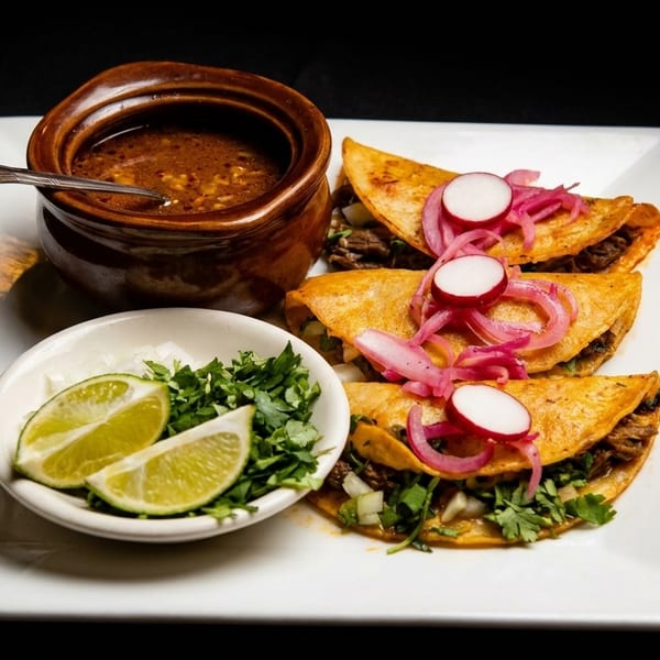 Three birria tacos with pickled red onions, radishes, cilantro, lime, and a side of consommé.