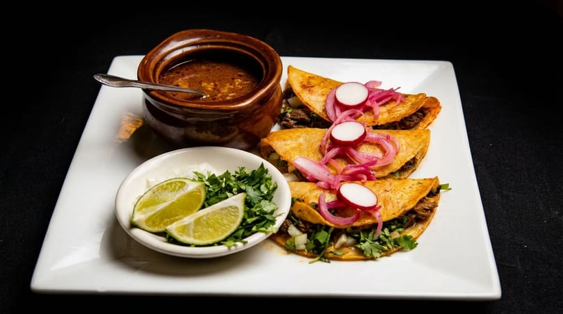 Three birria tacos with pickled red onions, radishes, cilantro, lime, and a side of consommé.
