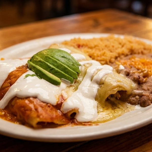 A plate of enchiladas with red and green sauce, sour cream, avocado, rice, and refried beans.
