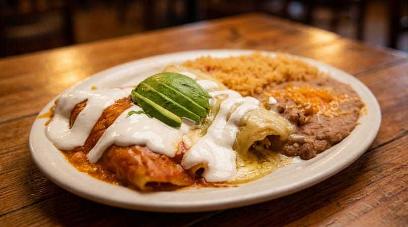 A plate of enchiladas with red and green sauce, sour cream, avocado, rice, and refried beans.