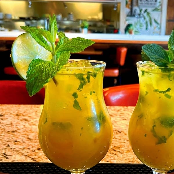 Two mojitos with lime and mint on a granite bar top, with a restaurant interior in the background.