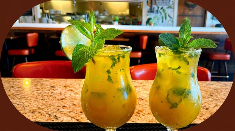 Two mojitos with lime and mint on a granite bar top, with a restaurant interior in the background.
