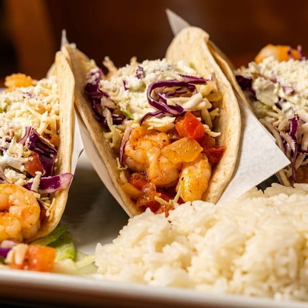Three shrimp tacos with cabbage, cheese, and tomatoes, served with a side of white rice.