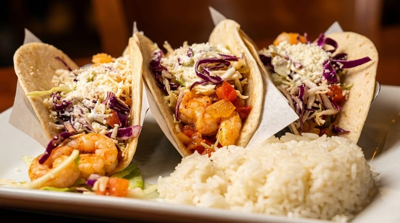 Three shrimp tacos with cabbage, cheese, and tomatoes, served with a side of white rice.