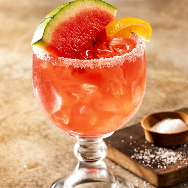 A vibrant red watermelon margarita, garnished with a watermelon slice and orange, sits on a rustic table.