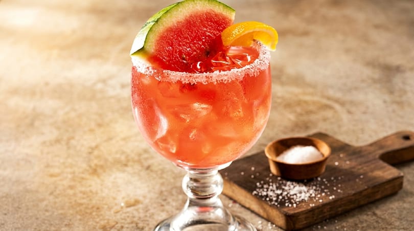 A vibrant red watermelon margarita, garnished with a watermelon slice and orange, sits on a rustic table.