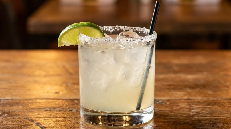 A margarita with a salted rim, lime wedge, and black straw on a wooden table.