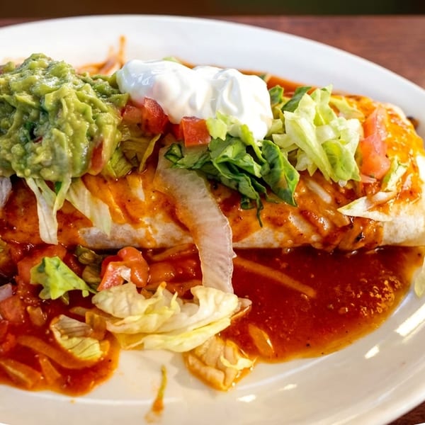 A large burrito covered in red sauce, guacamole, sour cream, and shredded lettuce on a white plate.