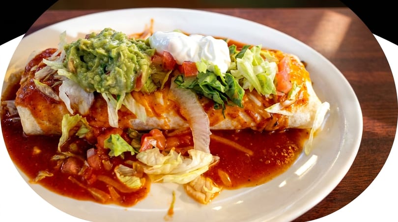 A large burrito covered in red sauce, guacamole, sour cream, and shredded lettuce on a white plate.